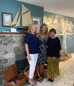 Lori Emery, Lynn Underhill and Debbie Ross stand together in front of the stone fireplace and tree of life recognition wall at the grand opening of the Ronald McDonald Family Retreat at Krantz Cottage.
