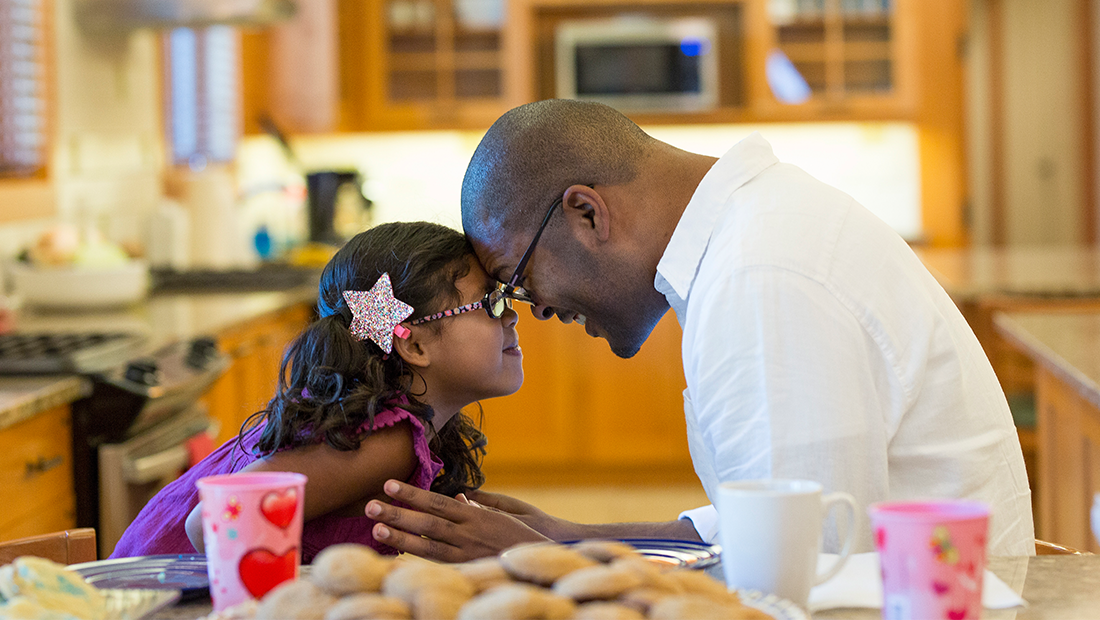 Father and daughter smiling forehead to forehead, behind a plate of cookies on the table in a Ronald McDonald House kitchen.