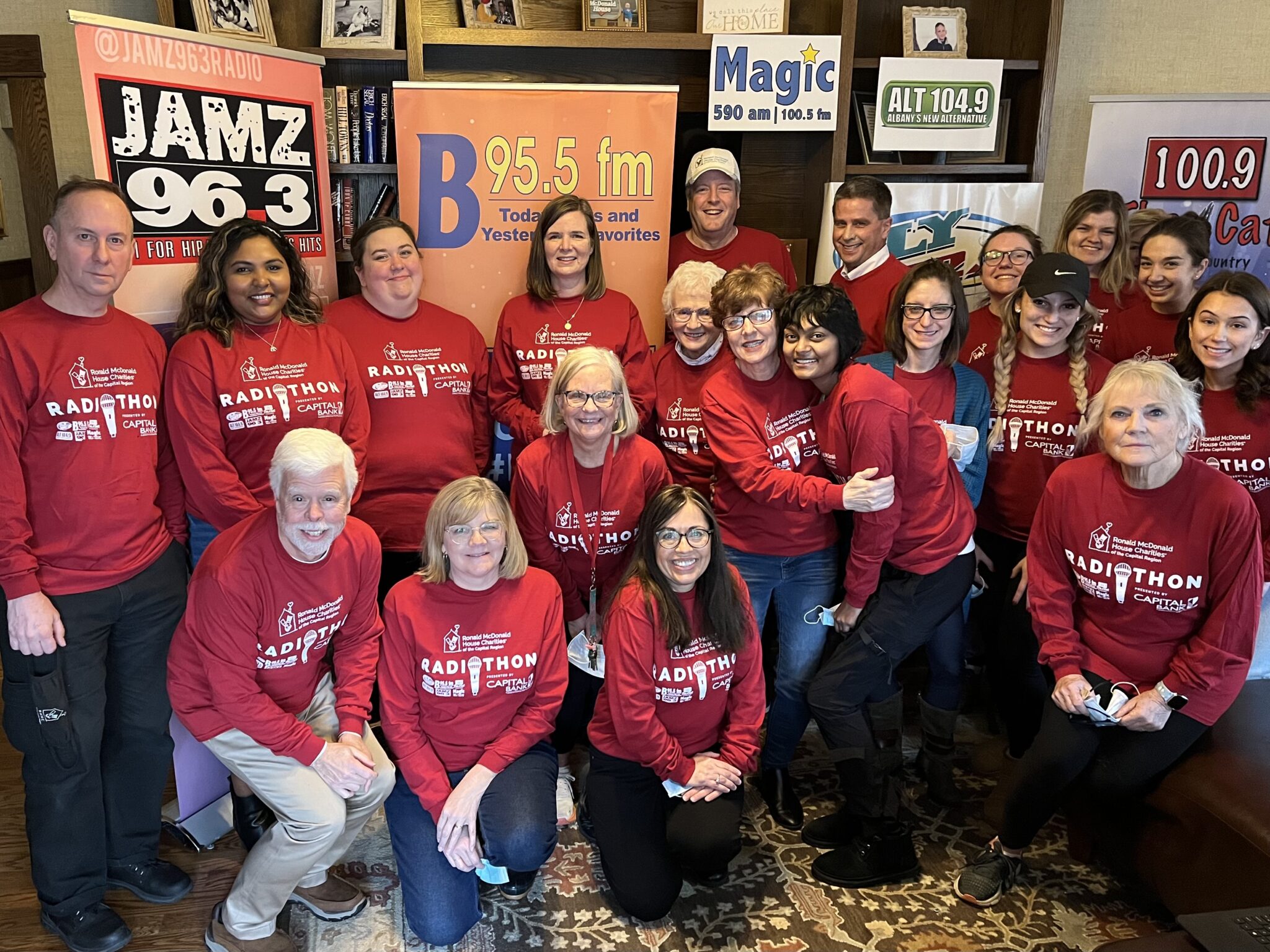 Large group of staff wearing red Radiothon t-shirts, surrounded by radio station banners in a large living room.
