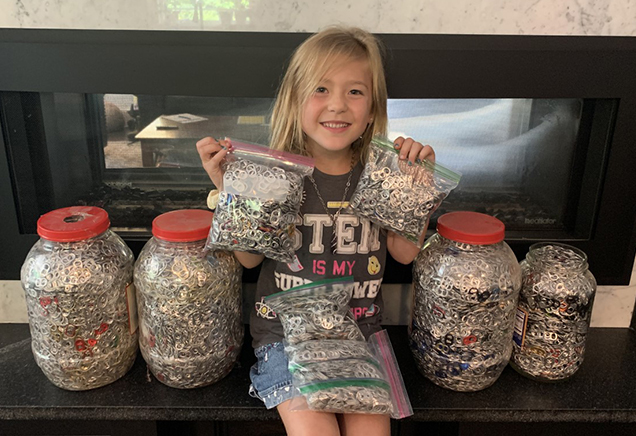 Young girl with blonde hair holds up clear bags full of pull-tabs, with even more in large containers all around her