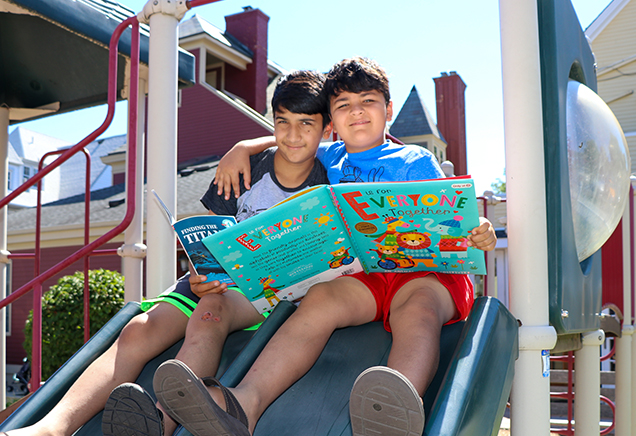 Two boys sitting on a playground slide with their arms around each other and books open on their laps