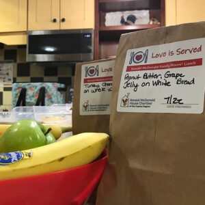 Close up of two brown-bag lunches with Love Is Served labels sitting on a counter next to a bowl of fruit