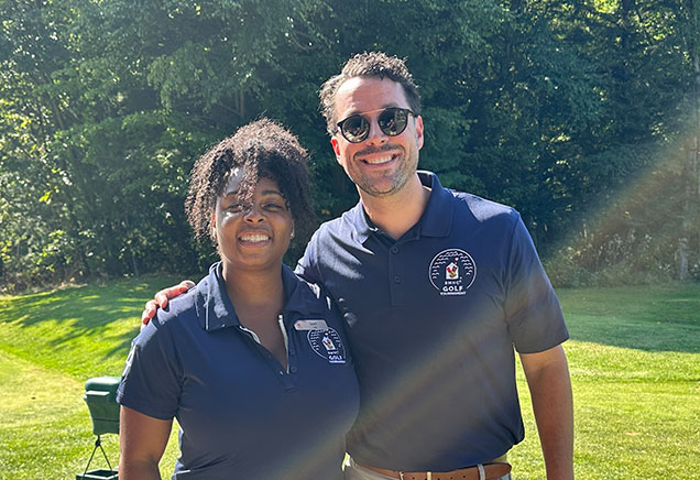 Two volunteers in navy blue polo shirts stand together on a golf course with the sun shining over them